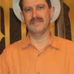 Man in an orange shirt and white hat standing in front of a patterned background.