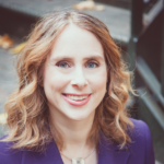 Smiling woman with wavy hair in a purple blazer, standing outside near stairs.