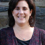 Smiling woman with dark hair, wearing a patterned top and necklace, standing in front of a stone wall.