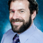 Smiling man with a beard wearing a purple shirt and tie against a blue background.