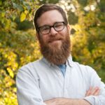 Man with a beard and glasses smiling outdoors with trees in the background.