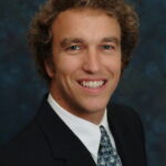 A person in a suit and tie smiles against a dark, textured background.