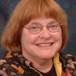 Smiling person with glasses and short red hair, wearing a patterned jacket against a dark background.