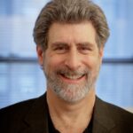 A smiling man with gray hair and a beard, wearing a dark blazer and shirt against a windowed background.