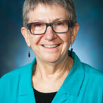 Person wearing glasses and a teal shirt, smiling against a dark, blurred background.