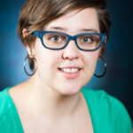 Person with short hair, blue glasses, and a green shirt smiles at the camera against a dark blue background.
