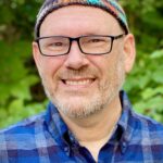 A man with glasses and a beanie smiles, wearing a blue plaid shirt in a green, outdoor setting.