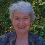 A person with short gray hair smiles in front of a leafy green background.