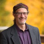 Man wearing glasses, a colorful hat, and a dark jacket, smiling with a blurred yellow background.