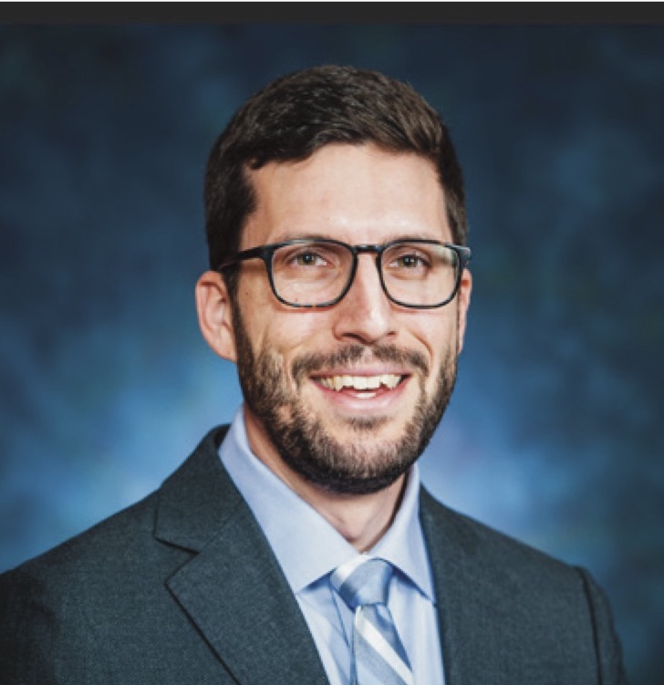 Rabbi Micah Weiss, wearing a jacket and tie, in front of a blue background.