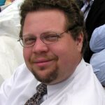 Smiling man with glasses and short curly hair, wearing a white shirt and patterned tie, sitting outdoors.