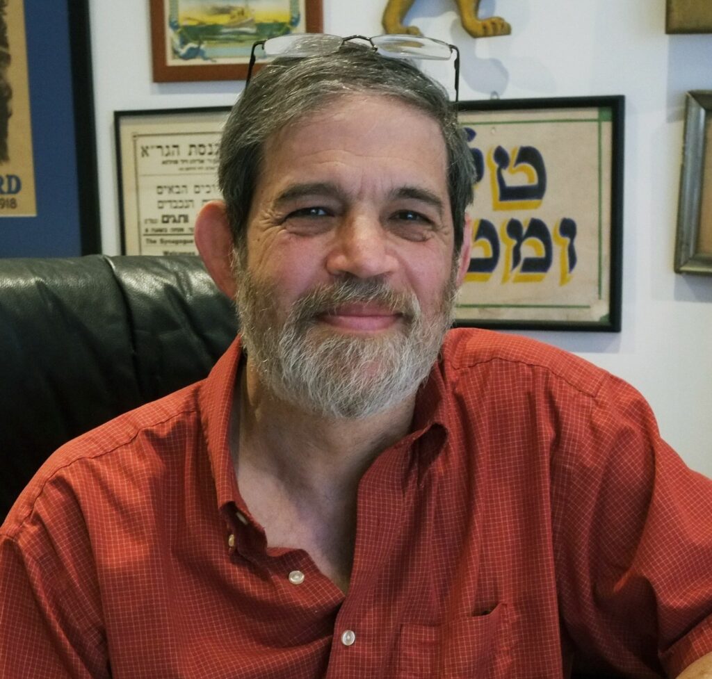 A smiling man with a beard wearing glasses on his head and a red shirt, sitting in front of framed artwork.