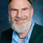 A smiling man with a beard wearing a colorful hat and a green jacket over a blue shirt.