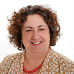 Smiling person with curly hair wearing a polka dot blouse and red necklace against a white background.