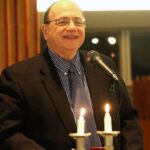 A man speaks at a podium with lit candles and an Israeli flag in the background.
