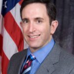 Smiling man in suit and colorful tie, standing in front of an American flag.