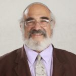 Smiling man with glasses, wearing a kippah, suit, and tie against a plain background.