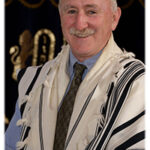 Smiling man in a suit and tallit stands in front of a decorative background.