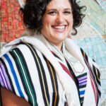Person smiling, wearing a striped shawl, standing in front of a colorful mosaic wall.