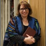 A person wearing glasses and a colorful shawl smiles while holding a book against a background of curtains.