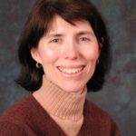 Smiling woman with short dark hair, wearing a brown sweater and turtleneck, against a textured backdrop.