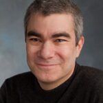 A person with short gray hair smiling, wearing a dark sweater against a neutral background.