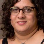 Person with curly hair and glasses, wearing a circular pendant necklace and a black top.