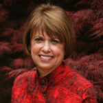 Smiling woman in a red patterned blouse with a background of deep red foliage.