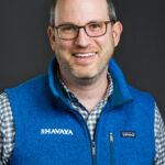 A smiling man wearing glasses, a blue fleece vest, and a checked shirt against a dark background.
