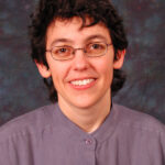 A person with short hair and glasses smiling, wearing a button-up shirt.