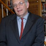 A man in a suit and red tie standing in a library, with bookshelves and wooden stairs in the background.