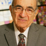 Elderly man in a suit and patterned tie, standing in front of a stained glass window.