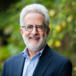 Smiling man in a suit with glasses and a beard, standing outdoors with greenery in the background.