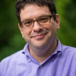 A person with glasses and a purple shirt smiles at the camera against a blurred green background.