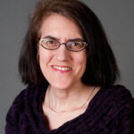 A person wearing glasses and a purple sweater smiles at the camera against a gray background.