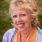 Smiling woman with curly blonde hair, wearing a colorful shawl and pearl necklace, outdoors.