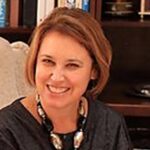 Smiling woman with short hair sitting indoors, wearing a dark top and a chunky necklace. Bookshelves in the background.