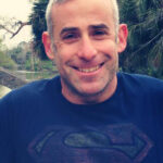 Smiling man wearing a Superman t-shirt with trees in the background.