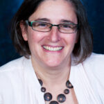 Smiling person with glasses and short hair wearing a necklace and light jacket against a dark blue background.