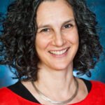 Smiling person with curly hair, wearing a red top and necklace, against a blue background.