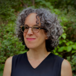 Person with curly hair and glasses smiling, wearing a sleeveless top, with a green leafy background.