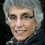 A person with short gray hair, glasses, and a scarf smiles at the camera.