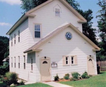 Two-story white house with beige doors and a circular window, surrounded by greenery on a sunny day.