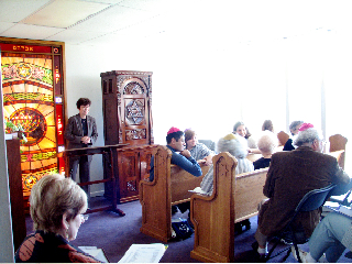 A person stands at a podium in a bright room with stained glass and attendees seated in wooden pews.