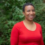 Smiling person in a red shirt standing in front of green foliage.