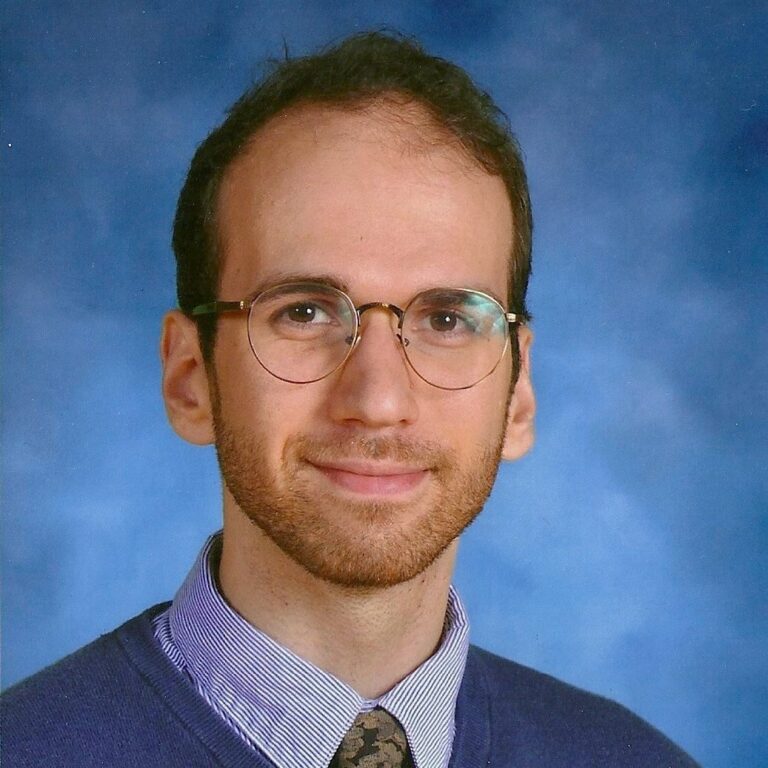 A person with glasses, wearing a tie, blue sweater, and collared shirt, smiles against a blue background.