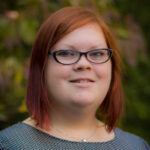 Person with red hair and glasses smiling, wearing a patterned top, with blurred greenery in the background.