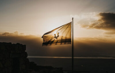 The Israeli flag waves against a dramatic sunset sky with dark clouds and a silhouette of stone ruins in the foreground.