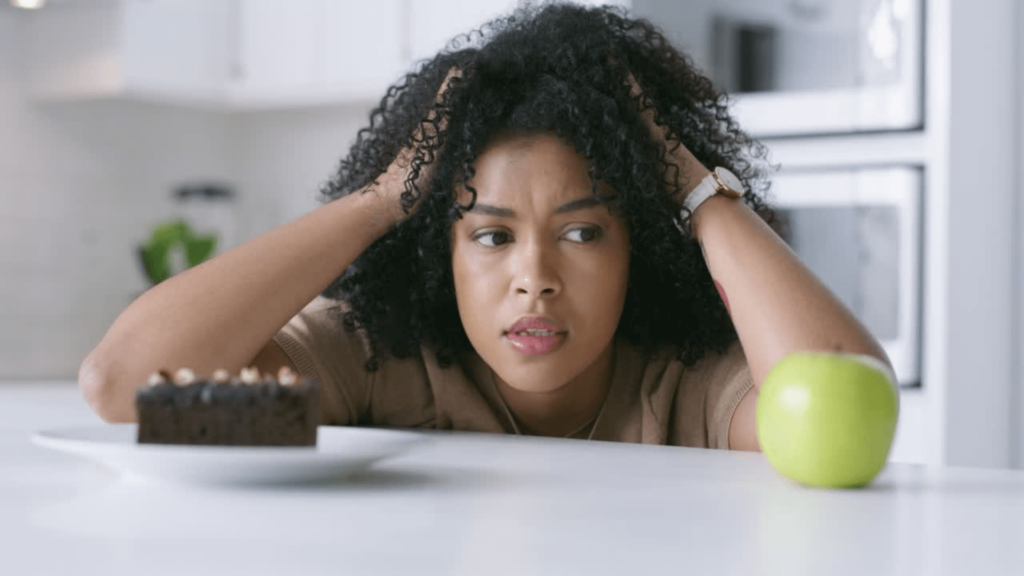 a Black woman in her kitchen looking confused at a piece of cake and an apple