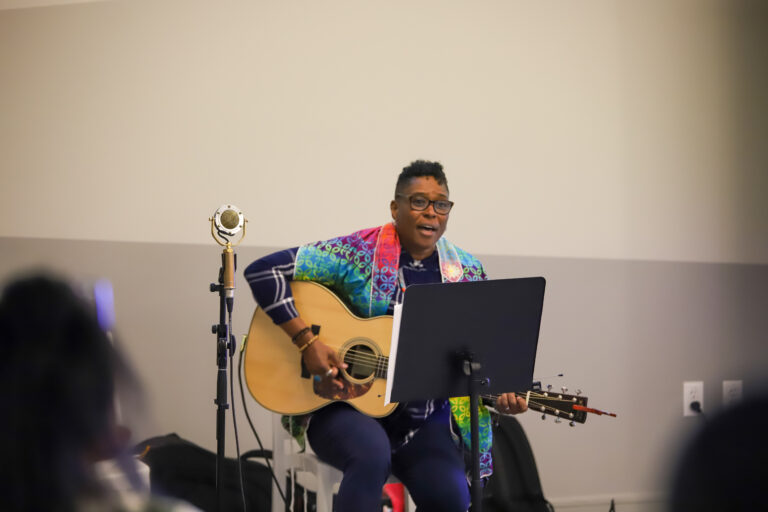 Rabbi Sandra Lawson playing guitar and singing at Juneteenth Shabbat wearing a rainbow colored tallit.